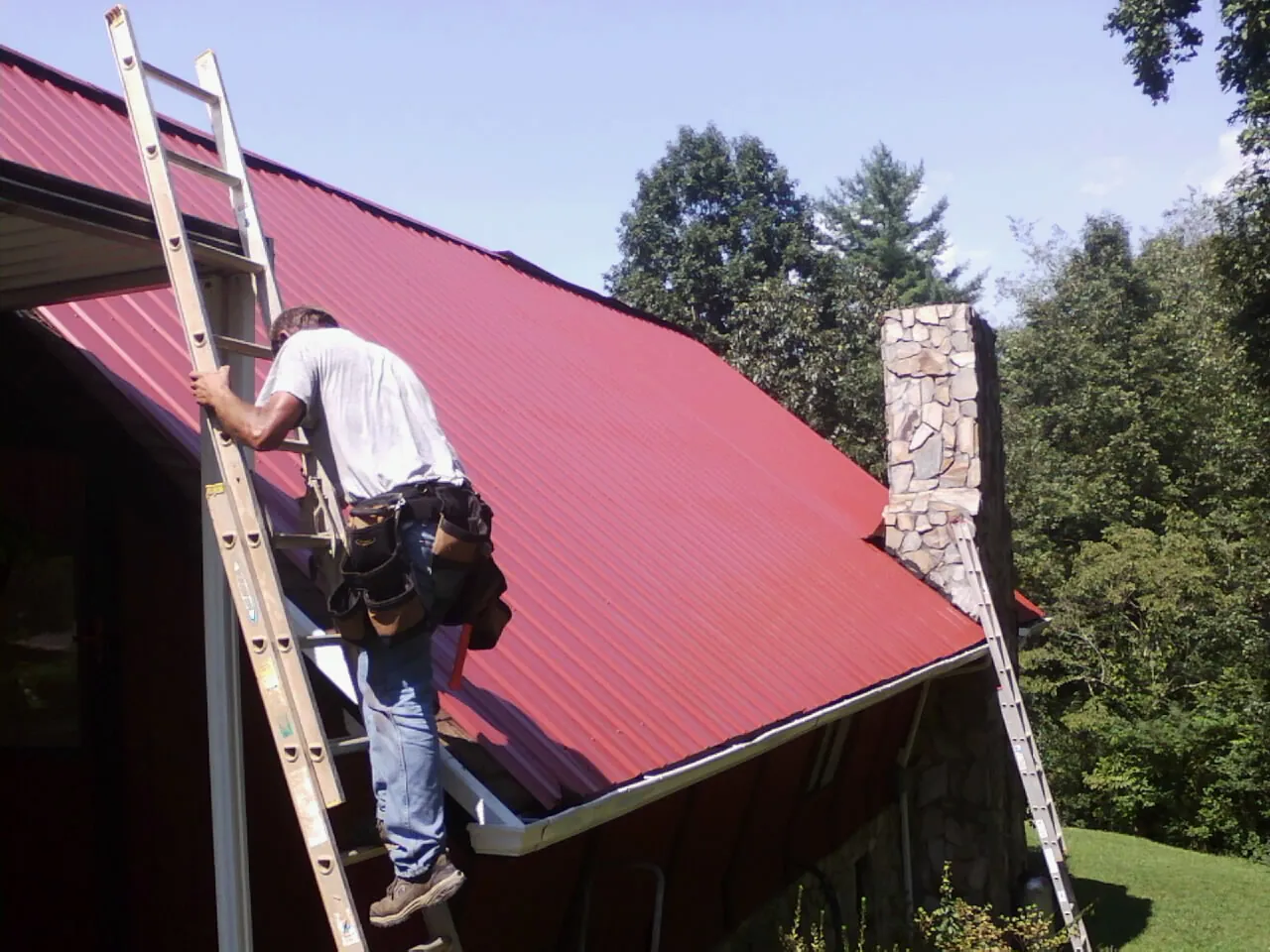 Roofer on a ladder beside a red metal roof and stone chimney — Soffit & Fascia Repair by Roofing Brothers in Clallam Bay